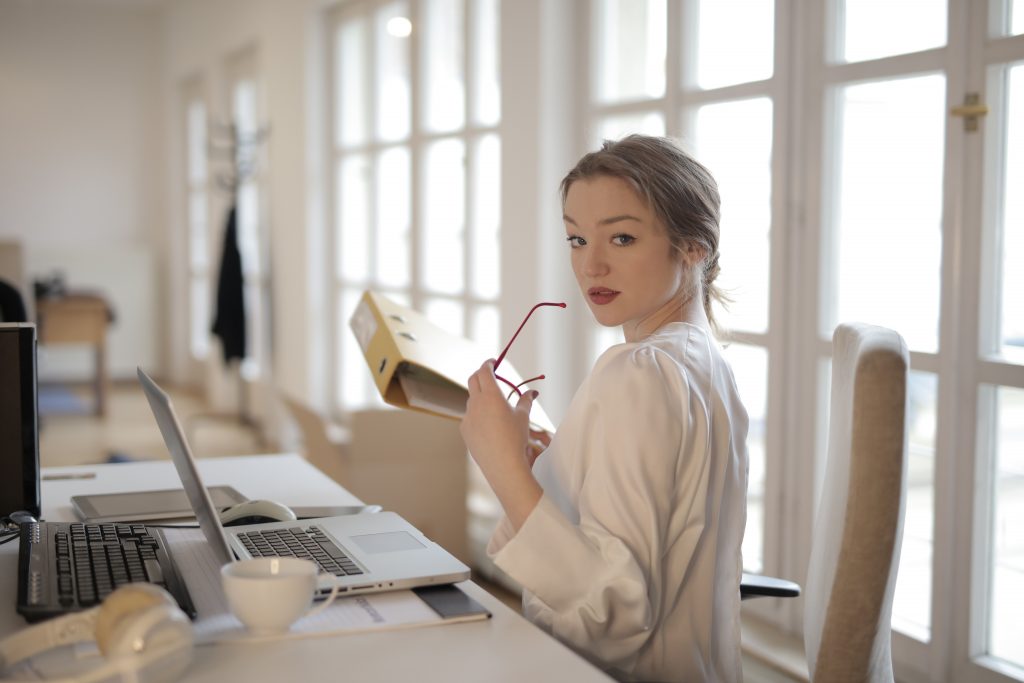 Back view of confident female entrepreneur in classy blouse sitting at table and doing paperwork in modern workplace while looking over shoulder
